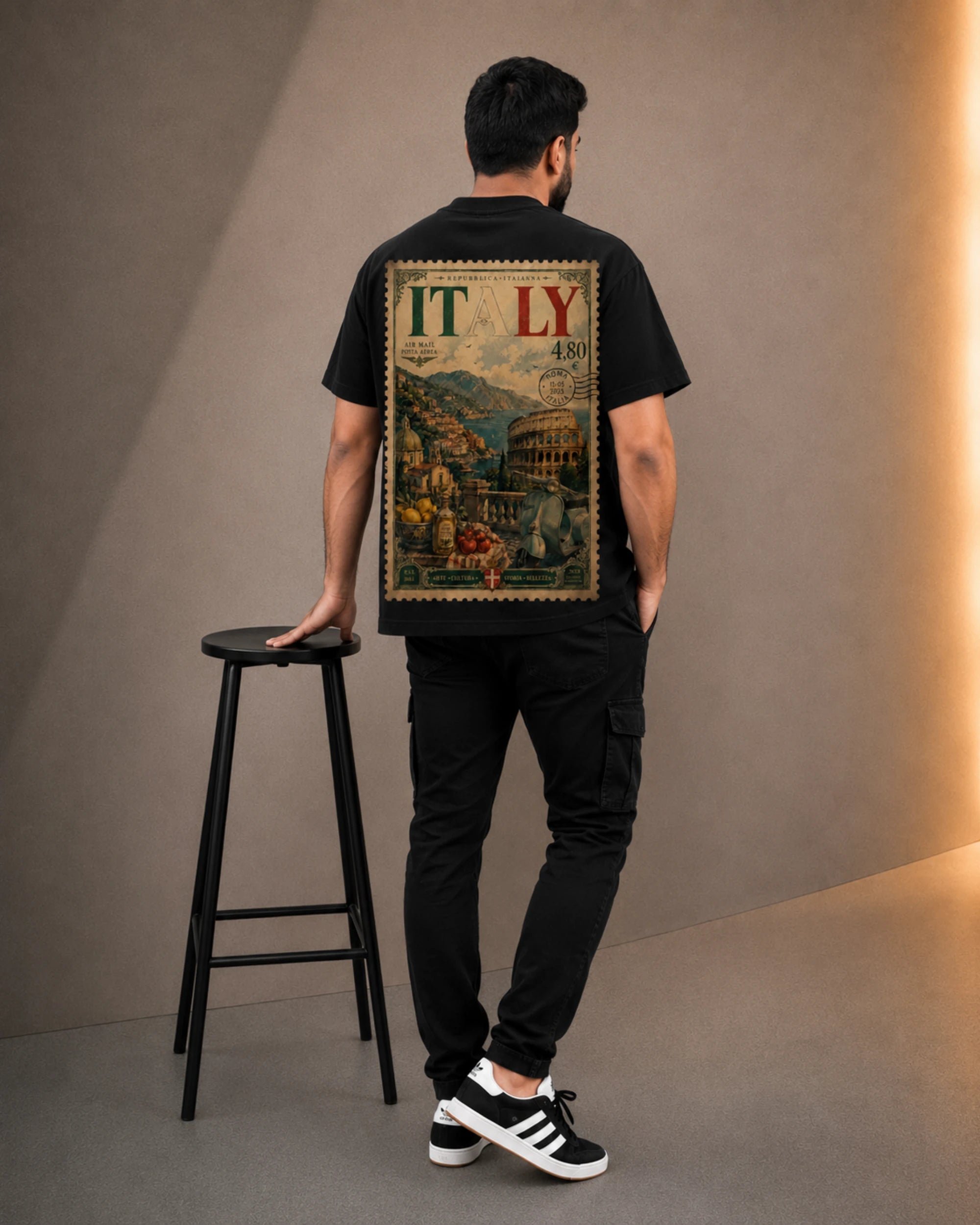 A man viewed from behind wearing a black t-shirt with a large vintage-style 'ITALY' travel poster graphic on the back, paired with black cargo pants and black-and-white sneakers, standing next to a black stool in a studio setting.
