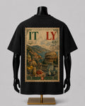 A black short-sleeve T-shirt displayed on a black mannequin torso with a gold stand, shown from the back. The shirt features a large rectangular vintage-style Italy travel-postmark graphic with the word ITALY, scenic artwork including the Colosseum and Mount Vesuvius, and postmark details on a light gray background.