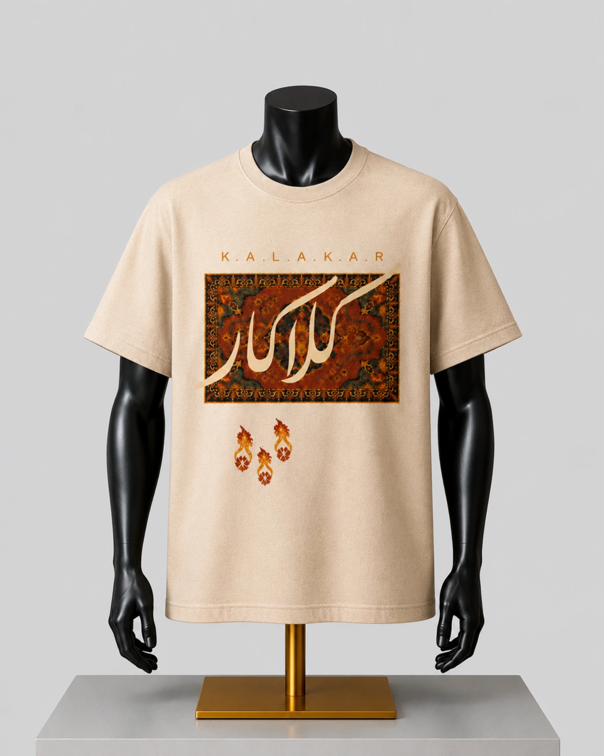 A beige short-sleeve t-shirt displayed on a black mannequin torso with a gold stand against a light gray background. The shirt features a graphic print with the word 'KALAKAR' in gold lettering above a rectangular patterned design with white Arabic calligraphy, and three small gold crescent moon icons below.