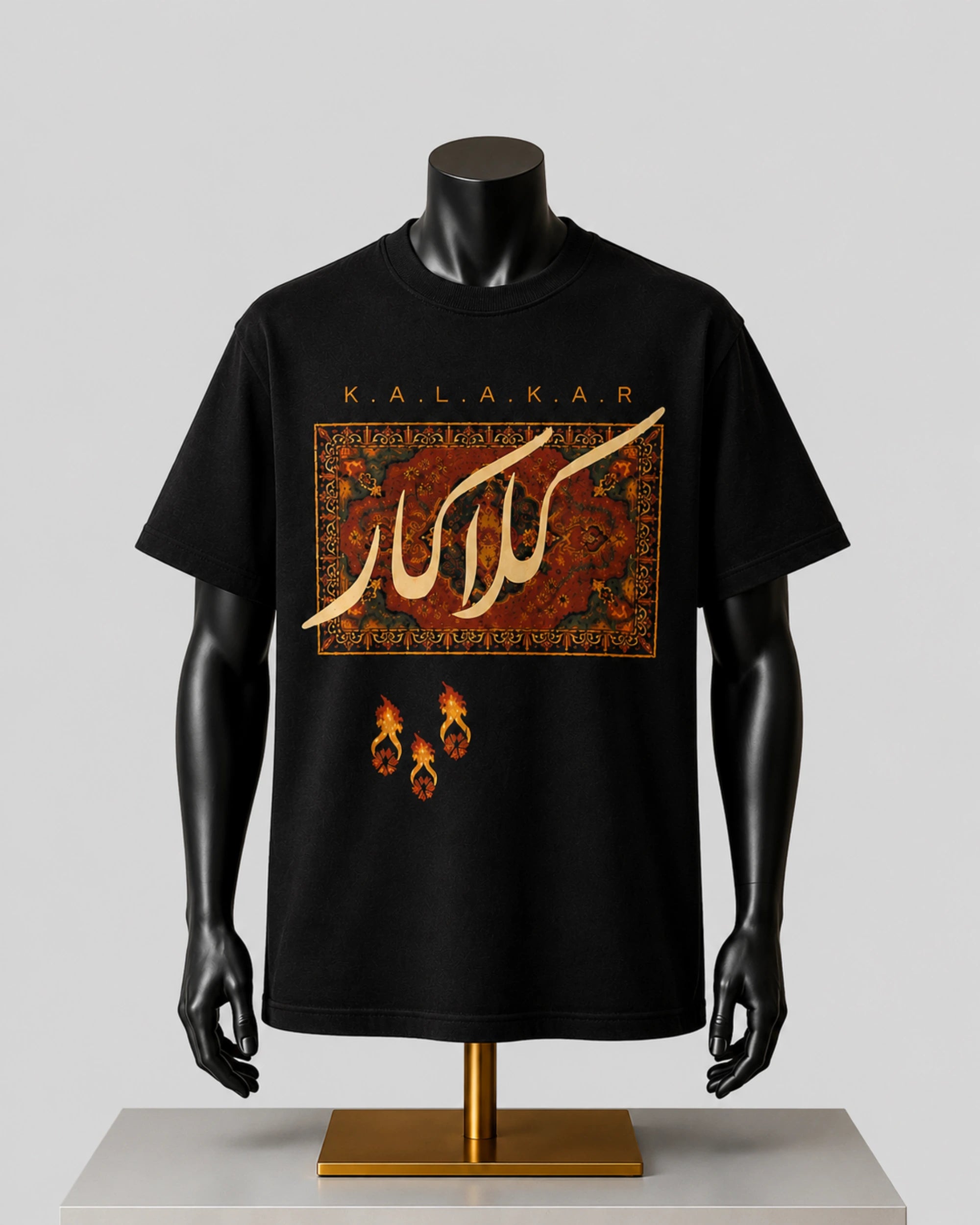 A black short-sleeve graphic T-shirt displayed on a black mannequin torso with arms. The shirt features a large rectangular red and gold patterned graphic with stylized white Arabic calligraphy, the word 'KALAKARD' above it, and three small flame-like motifs below.