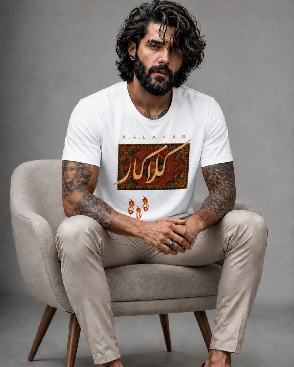 A man with a beard and curly hair sits in a gray armchair wearing a white graphic t-shirt with Arabic calligraphy and a matching pair of dangling earrings. The t-shirt features a rectangular brown and gold pattern with the word 'KALAM' printed above it.