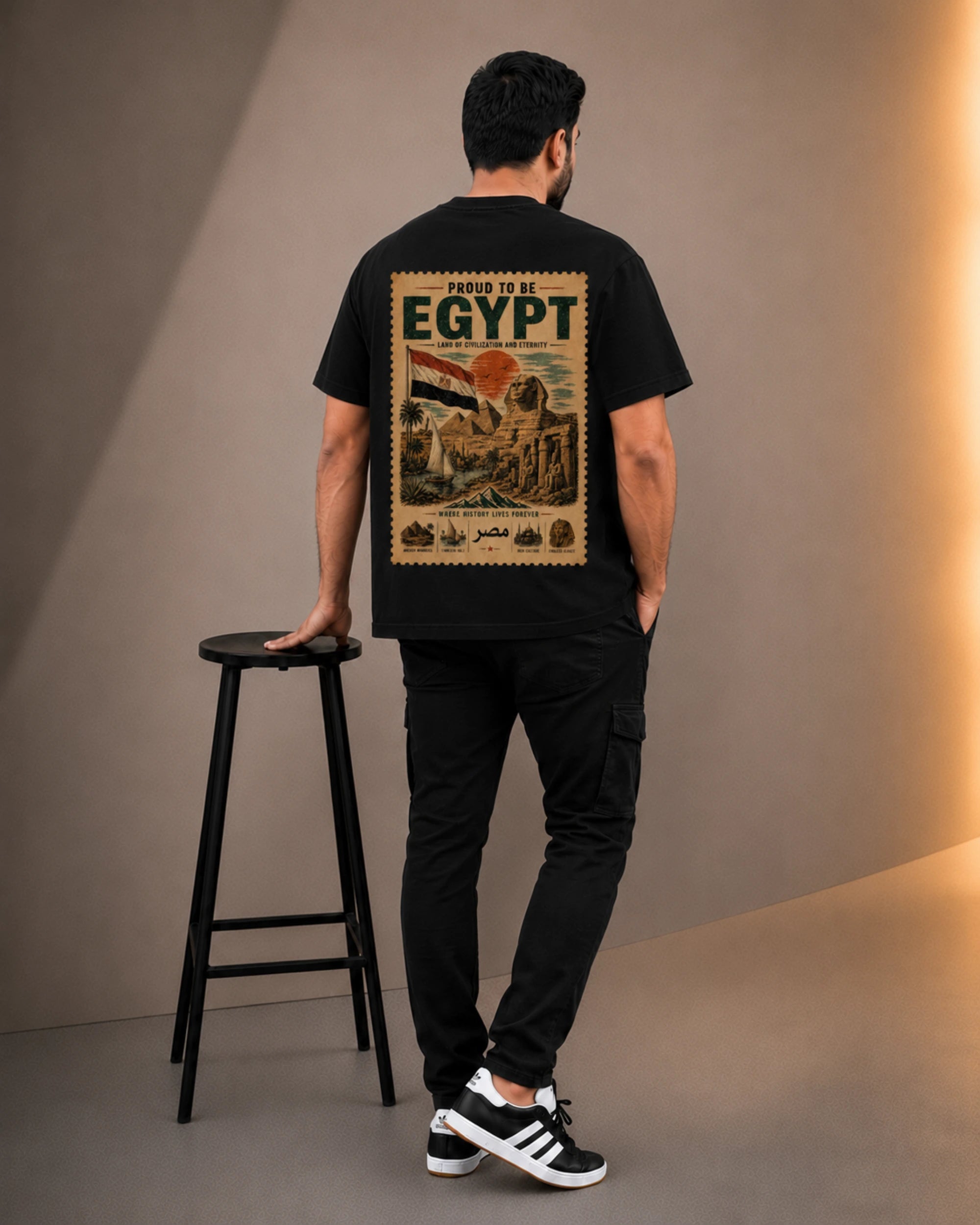 A man stands with his back to the camera wearing a black t-shirt with a large vintage-style Egyptian travel poster graphic on the back, paired with black cargo pants and black-and-white sneakers. He is standing next to a black metal stool in a studio setting with warm side lighting.