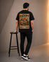 A man stands with his back to the camera wearing a black t-shirt with a large vintage-style Egyptian travel poster graphic on the back, paired with black cargo pants and black-and-white sneakers. He is standing next to a black metal stool in a studio setting with warm side lighting.
