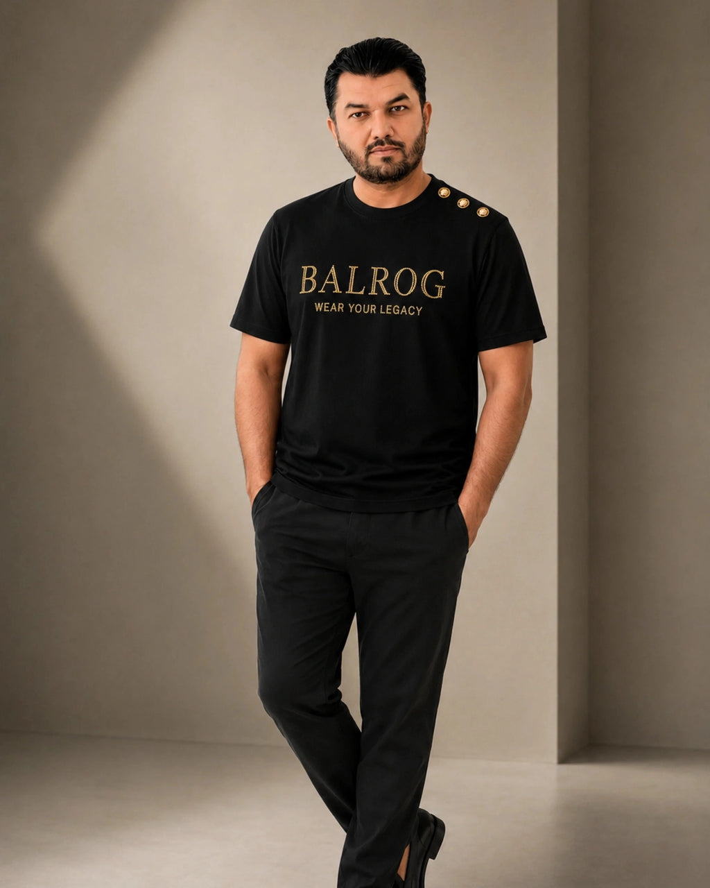 A man modeling a black Balrog t-shirt with gold lettering and three gold buttons on the shoulder, paired with black trousers, in a minimalist studio setting.