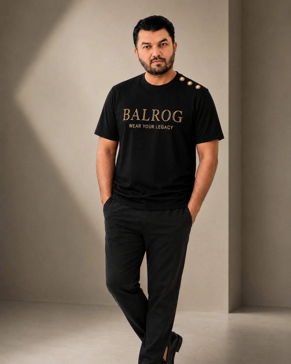 A man modeling a black Balrog t-shirt with gold lettering and three gold buttons on the shoulder, paired with black trousers, in a minimalist studio setting.