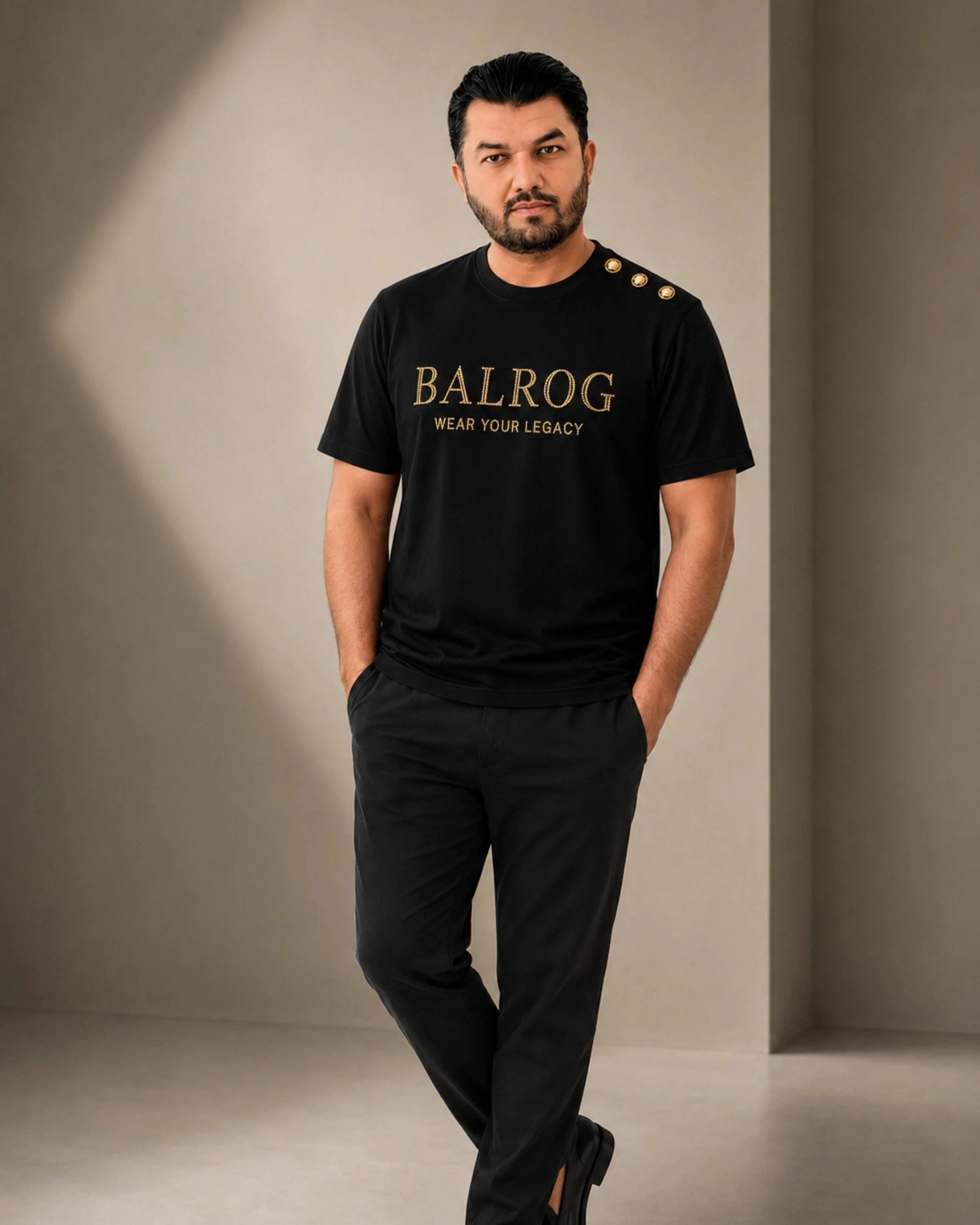 A man modeling a black Balrog t-shirt with gold lettering and three gold buttons on the shoulder, paired with black trousers, in a minimalist studio setting.