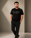 A man modeling a black Balrog t-shirt with gold lettering and three gold buttons on the shoulder, paired with black trousers, in a minimalist studio setting.
