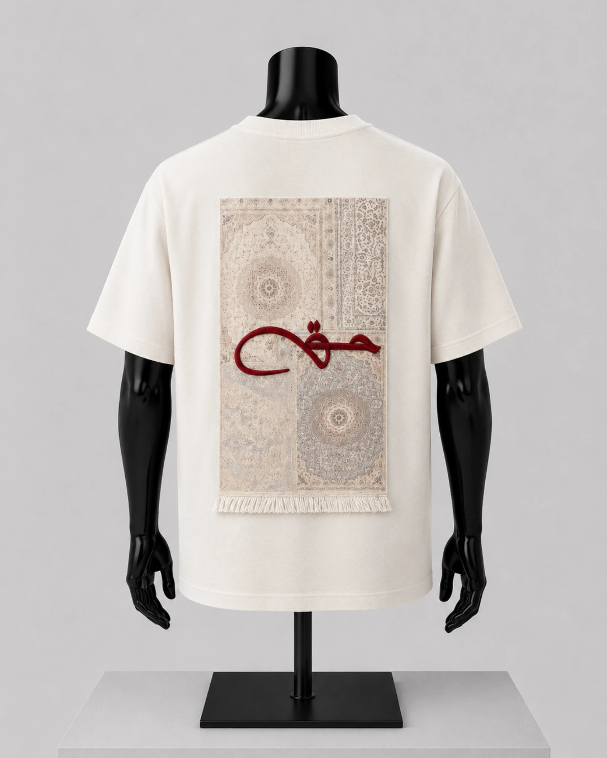 An off-white short-sleeve T-shirt displayed on a black mannequin torso, showing the back side with a large rectangular beige rug print and a red Arabic calligraphy graphic across the center.