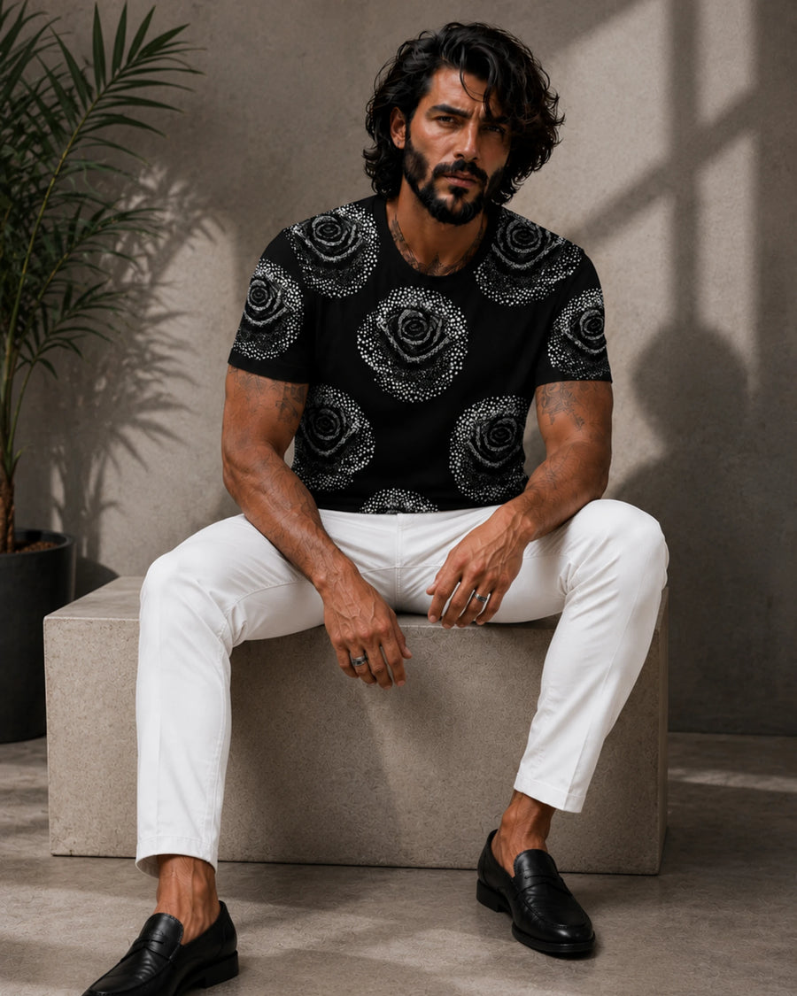 A man wearing a black short-sleeve t-shirt with a white rose pattern, white trousers, and black loafers, sitting on a concrete block in a modern indoor setting.