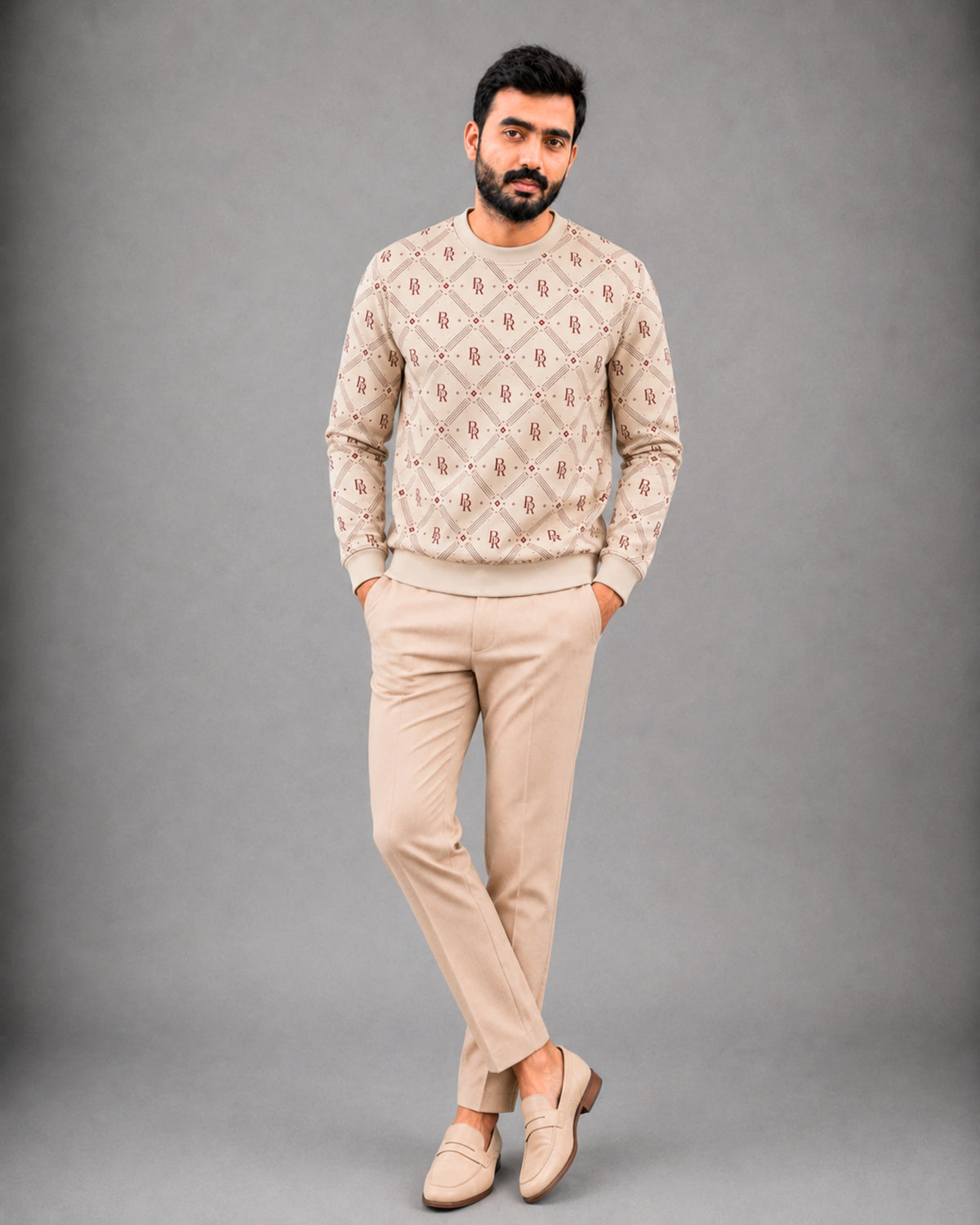 A man modeling a beige crewneck sweater with a brown geometric pattern, paired with beige trousers and matching loafers, against a solid gray studio background.