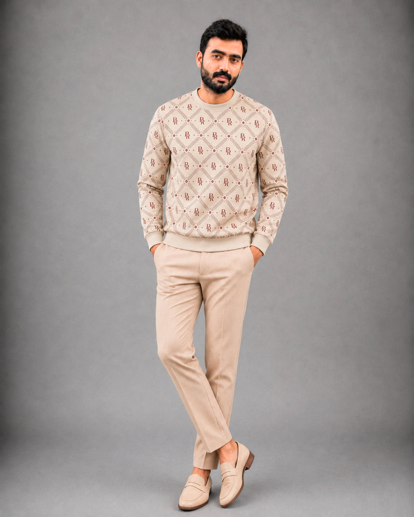 A man modeling a beige crewneck sweater with a brown geometric pattern, paired with beige trousers and matching loafers, against a solid gray studio background.