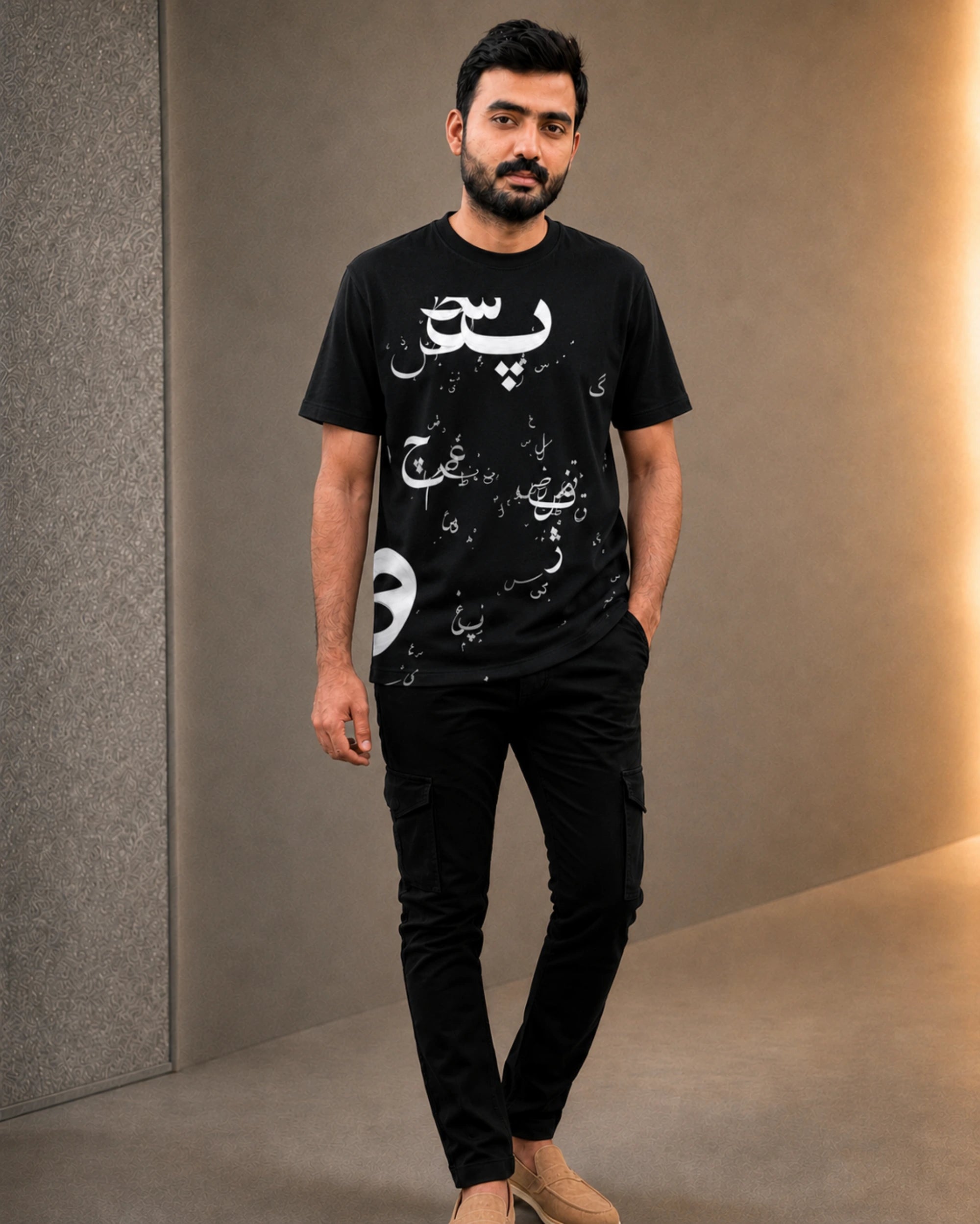 A man wearing a black short-sleeve t-shirt with white Arabic calligraphy and a distressed graphic print, paired with black cargo pants and beige shoes, standing in a modern indoor setting with a warm-toned wall.