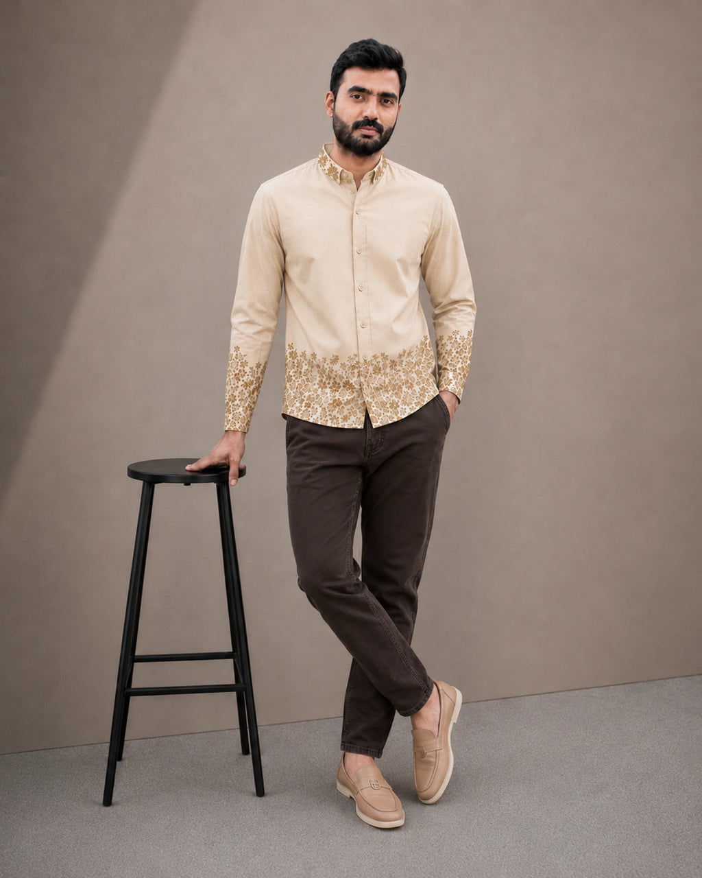 A man modeling a beige long-sleeve kurta with gold embroidery on the hem and cuffs, paired with dark brown trousers and beige loafers, standing next to a black stool against a neutral studio backdrop.