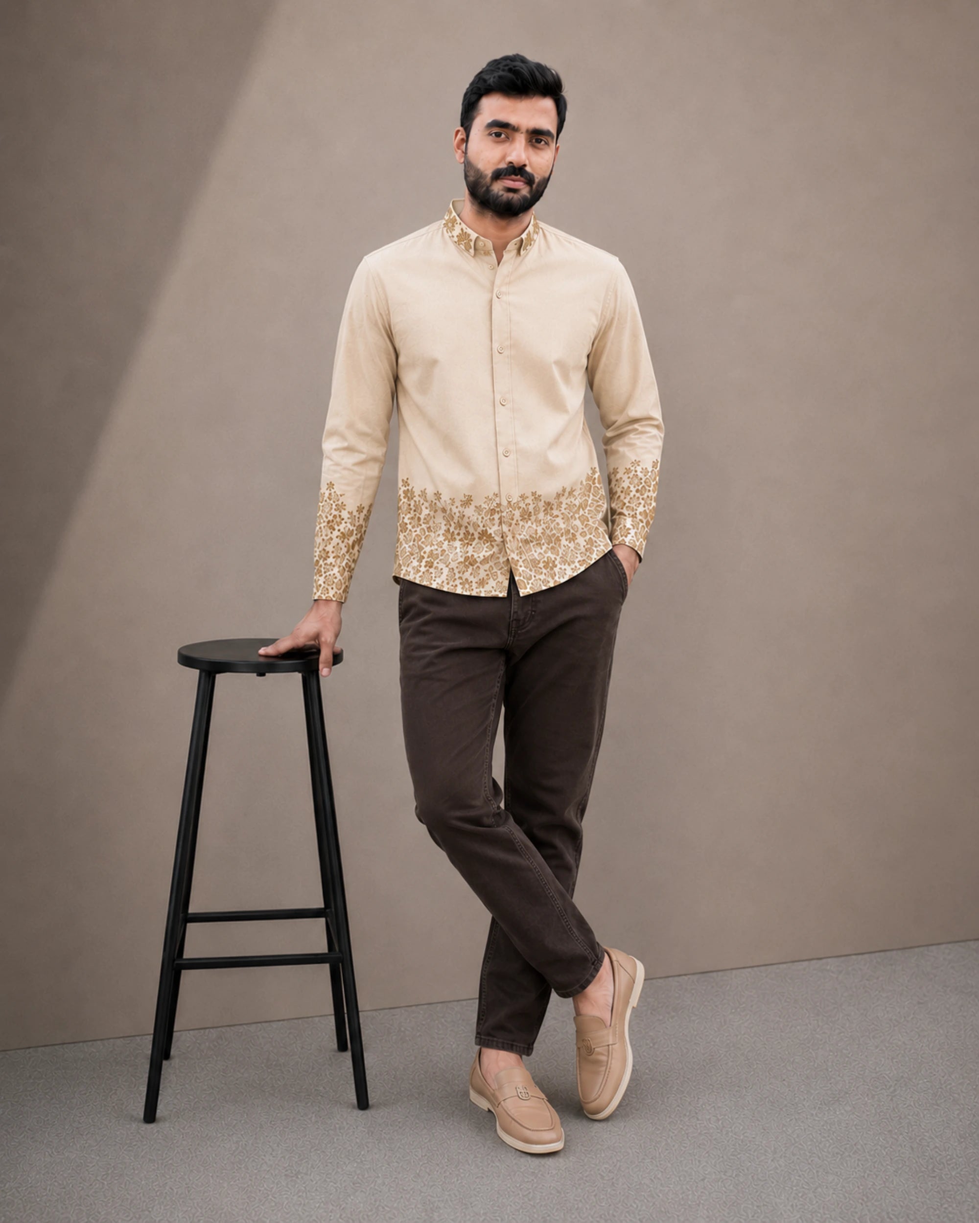 A man modeling a beige long-sleeve kurta with gold embroidery on the hem and cuffs, paired with dark brown trousers and beige loafers, standing next to a black stool against a neutral studio backdrop.