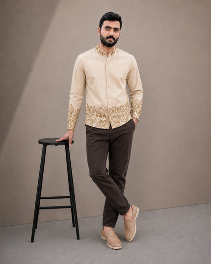 A man modeling a beige long-sleeve kurta with gold embroidery on the hem and cuffs, paired with dark brown trousers and beige loafers, standing next to a black stool against a neutral studio backdrop.