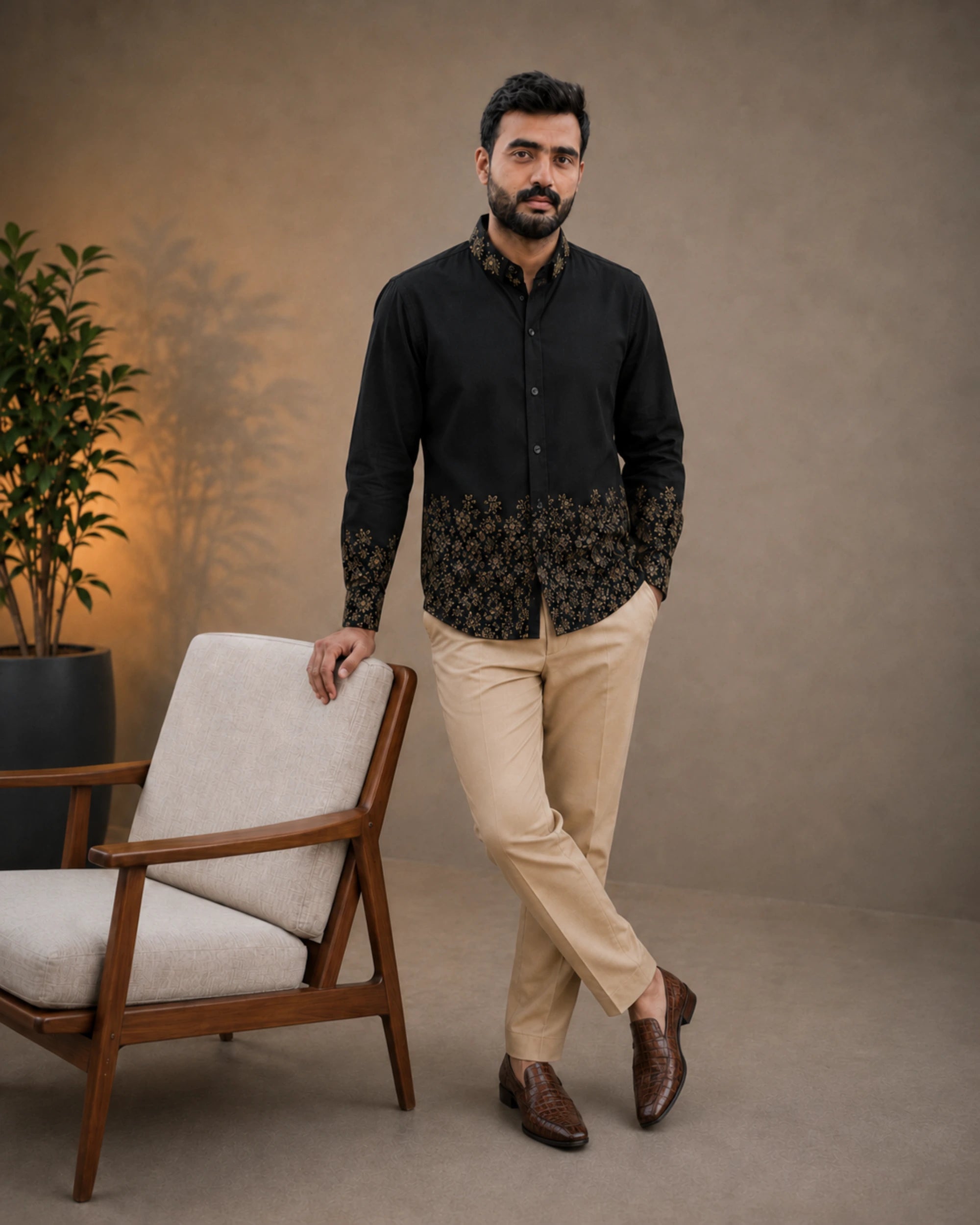 A man wearing a black long-sleeve button-up shirt with a beige and black floral patterned hem and cuffs, paired with beige trousers and brown loafers, standing next to a modern wooden armchair in a studio setting.