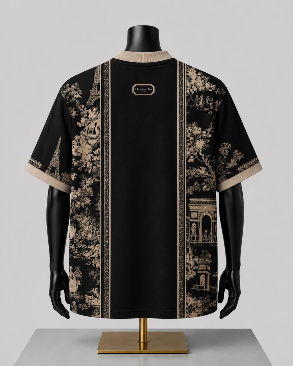 A black short-sleeve t-shirt with beige toile de Jouy prints on the sides and a central vertical stripe, displayed on a black mannequin torso with a gold stand against a light gray background.