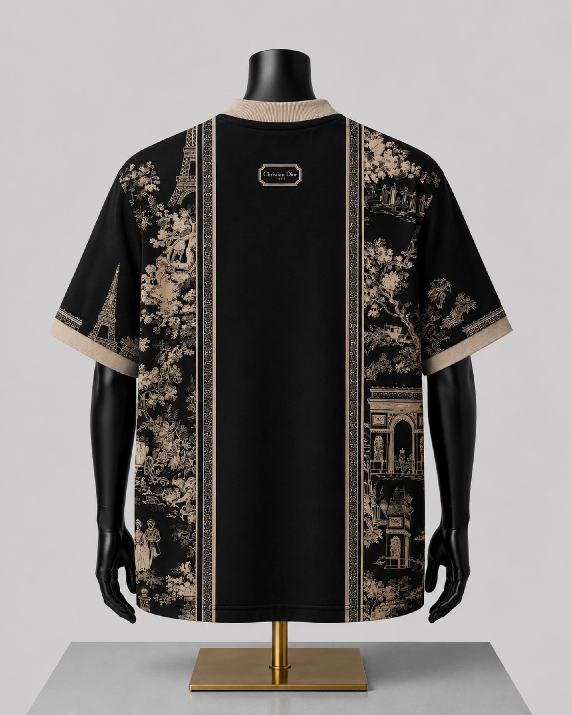 A black short-sleeve t-shirt with beige toile de Jouy prints on the sides and a central vertical stripe, displayed on a black mannequin torso with a gold stand against a light gray background.