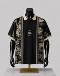 A black short-sleeve polo shirt with beige collar and sleeve trim, featuring beige scenic architectural and floral prints on the sides and a central gold-toned chest graphic, displayed on a black mannequin torso over a gold stand against a light gray background.