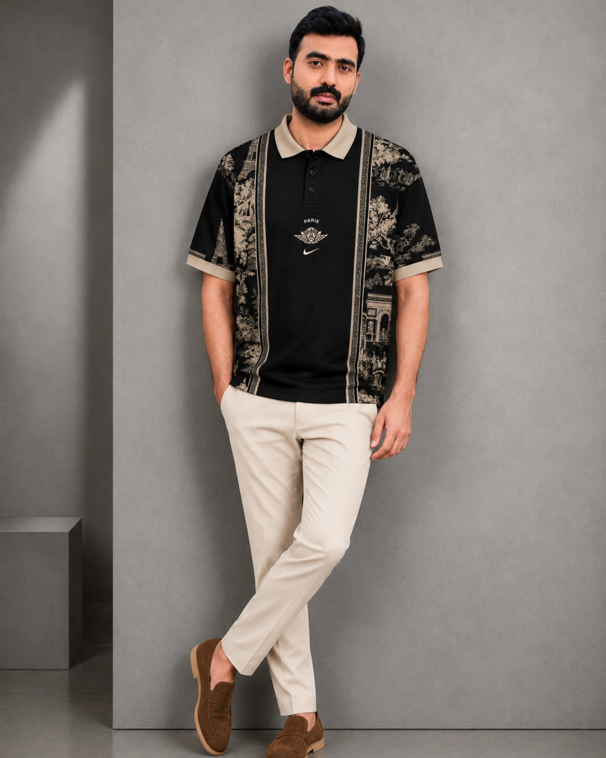 A man wearing a black short-sleeve polo shirt with beige floral-patterned panels on the sleeves and collar, paired with beige trousers and brown loafers, standing against a gray wall.
