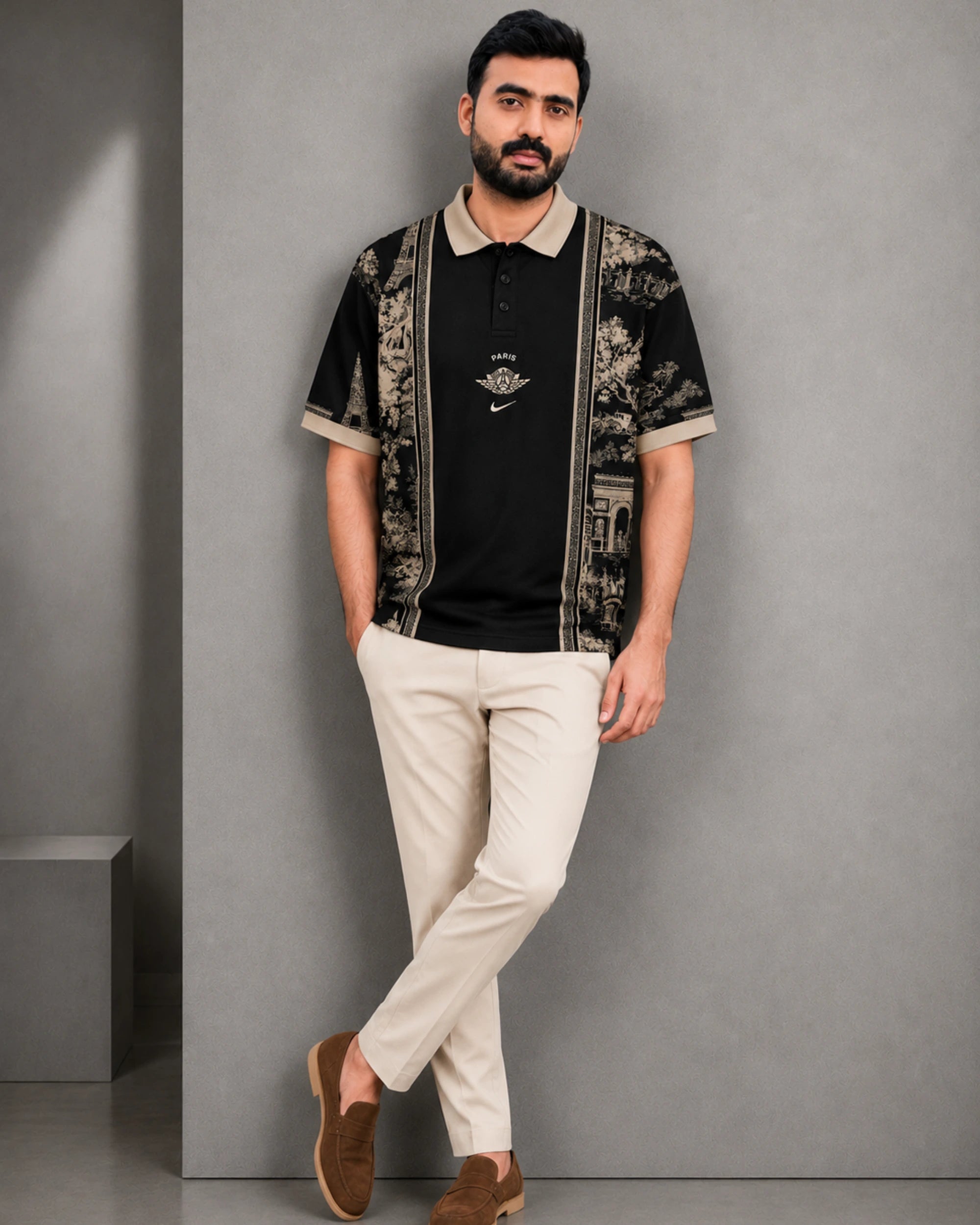 A man wearing a black short-sleeve polo shirt with beige floral-patterned panels on the sleeves and collar, paired with beige trousers and brown loafers, standing against a gray wall.