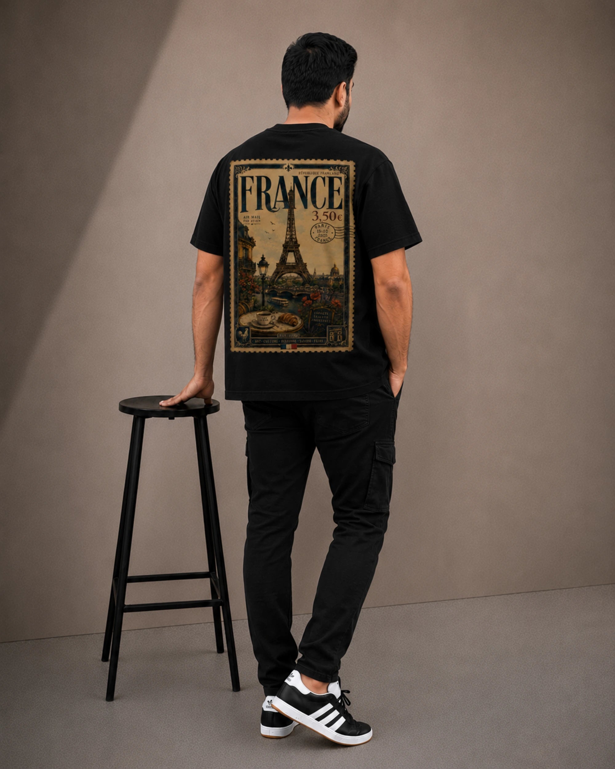 A man stands with his back to the camera wearing a black t-shirt with a large vintage-style 'FRANCE 1900' graphic print on the back, paired with black cargo pants and black-and-white sneakers. He is posed next to a black metal stool in front of a neutral gray studio backdrop.