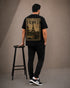 A man stands with his back to the camera wearing a black t-shirt with a large vintage-style 'FRANCE 1900' graphic print on the back, paired with black cargo pants and black-and-white sneakers. He is posed next to a black metal stool in front of a neutral gray studio backdrop.