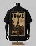 A black short-sleeve T-shirt displayed on a black mannequin torso with a gold stand, shown from the back. The shirt features a large vintage-style France postage stamp graphic with the Eiffel Tower, postmark details, and cafe imagery.