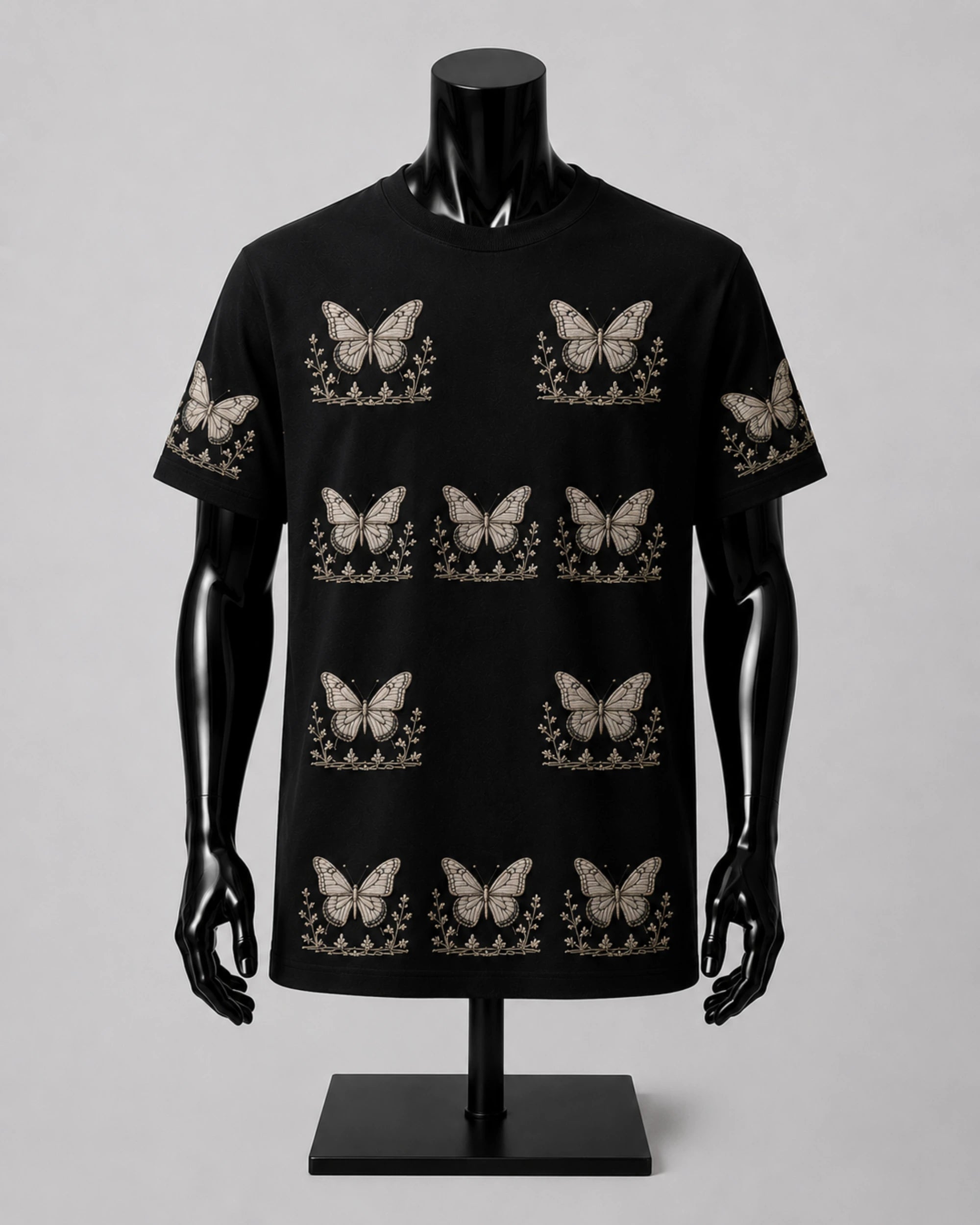 A black short-sleeve crewneck t-shirt displayed on a black mannequin torso. The shirt features an all-over repeating pattern of beige butterflies with floral details underneath.