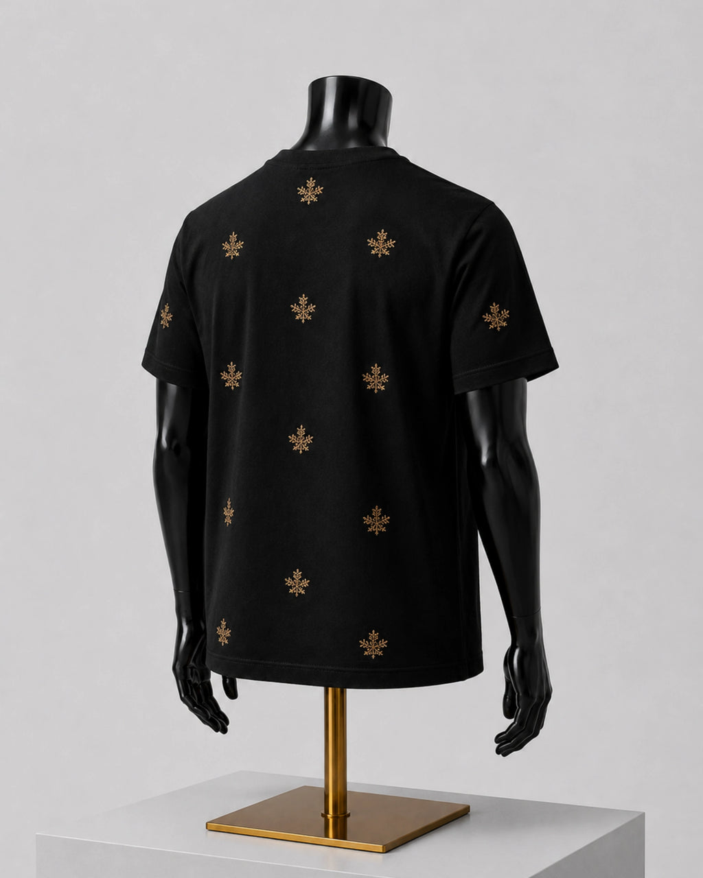 A black short-sleeve T-shirt displayed on a black mannequin torso with a gold stand, shown from the back. The shirt features a repeating pattern of small gold fleur-de-lis motifs, photographed against a neutral light gray background.