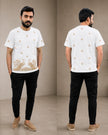 A man modeling a white short-sleeve t-shirt with gold peacock embroidery on the front and small gold motifs on the back, paired with black cargo pants and beige slip-on shoes, shown in front and back views against a neutral studio backdrop.
