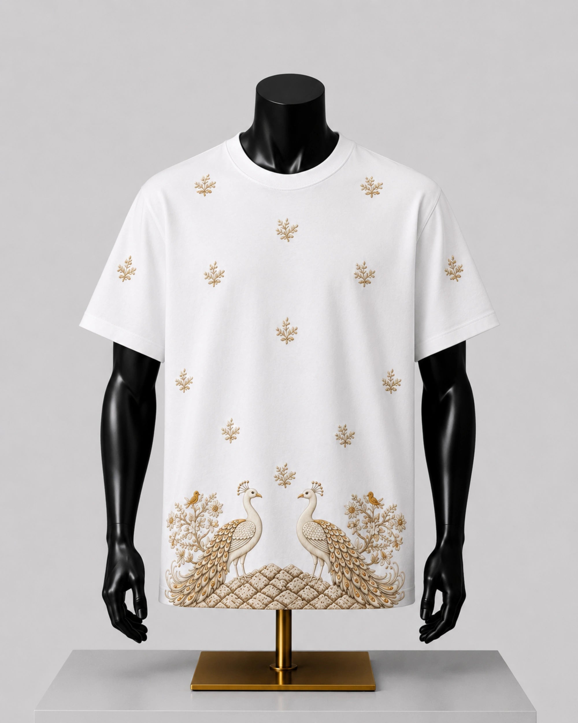 White short-sleeve t-shirt displayed on a black mannequin torso, featuring scattered gold floral motifs and a large peacock and bird pattern across the lower hem, set against a light gray background.