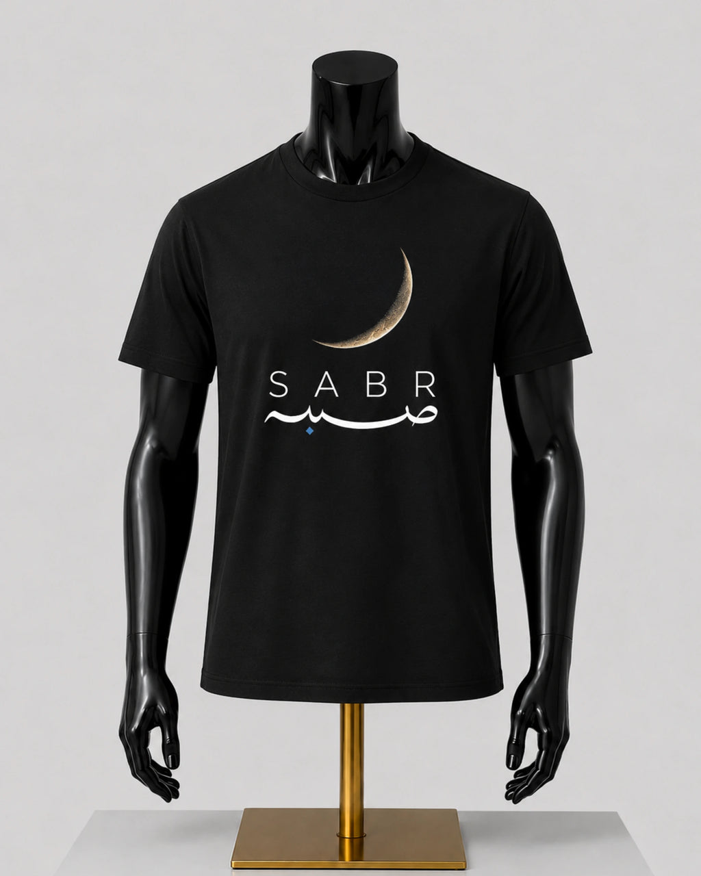 A black short-sleeve t-shirt displayed on a black mannequin torso with a gold stand against a light gray studio background. The shirt features a crescent moon graphic above the word 'SABR' in white, with white Arabic calligraphy and a small blue star accent.