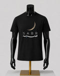A black short-sleeve t-shirt displayed on a black mannequin torso with a gold stand against a light gray studio background. The shirt features a crescent moon graphic above the word 'SABR' in white, with white Arabic calligraphy and a small blue star accent.