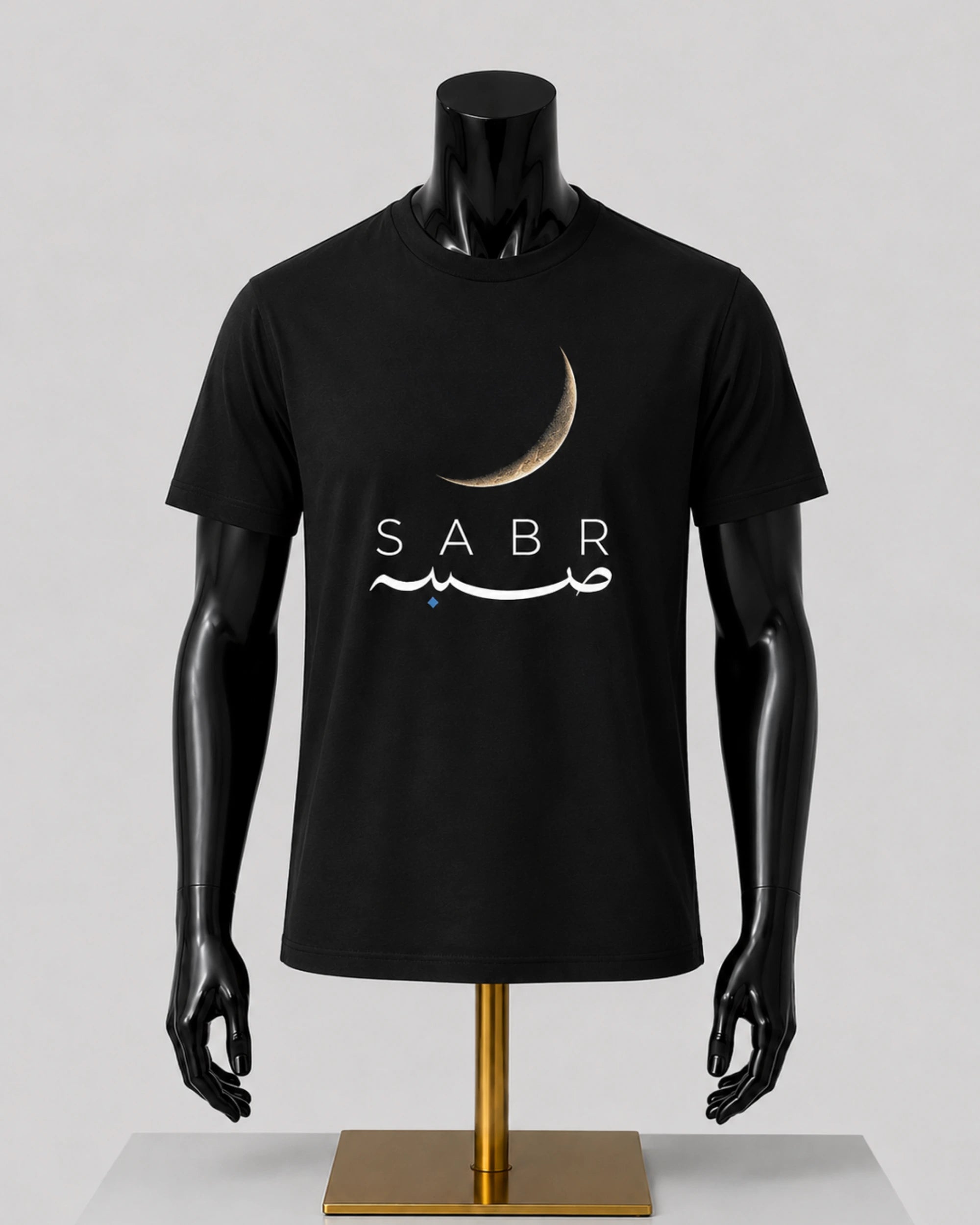 A black short-sleeve t-shirt displayed on a black mannequin torso with a gold stand against a light gray studio background. The shirt features a crescent moon graphic above the word 'SABR' in white, with white Arabic calligraphy and a small blue star accent.