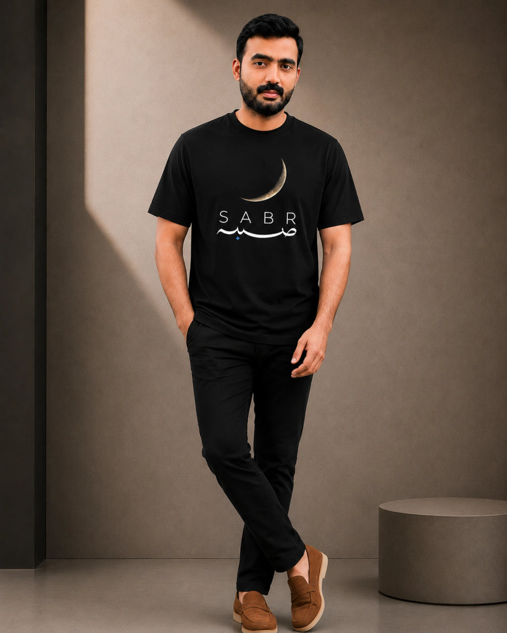 A man wearing a black crew-neck t-shirt with a white crescent moon and 'SABR' graphic, paired with black pants and brown slip-on shoes, standing against a textured gray wall.