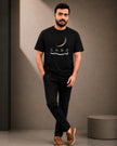 A man wearing a black crew-neck t-shirt with a white crescent moon and 'SABR' graphic, paired with black pants and brown slip-on shoes, standing against a textured gray wall.
