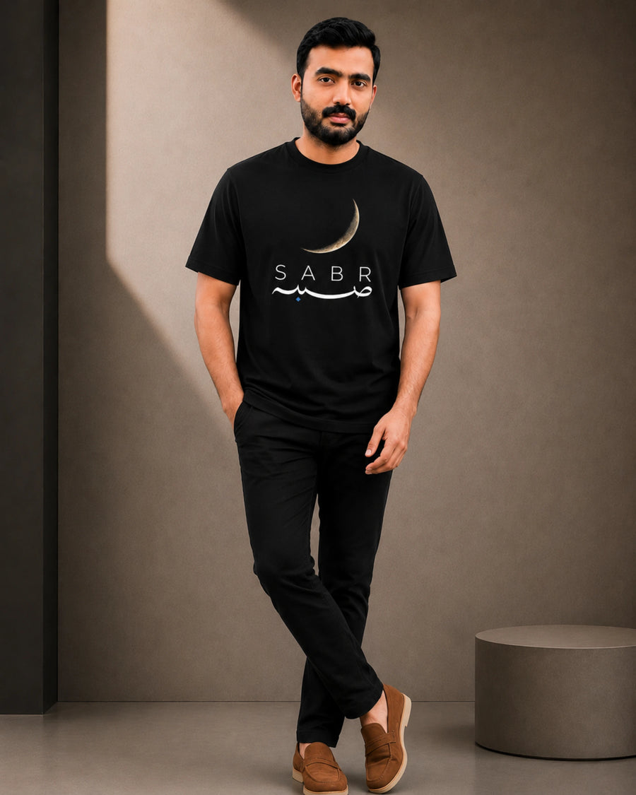 A man wearing a black crew-neck t-shirt with a white crescent moon and 'SABR' graphic, paired with black pants and brown slip-on shoes, standing against a textured gray wall.