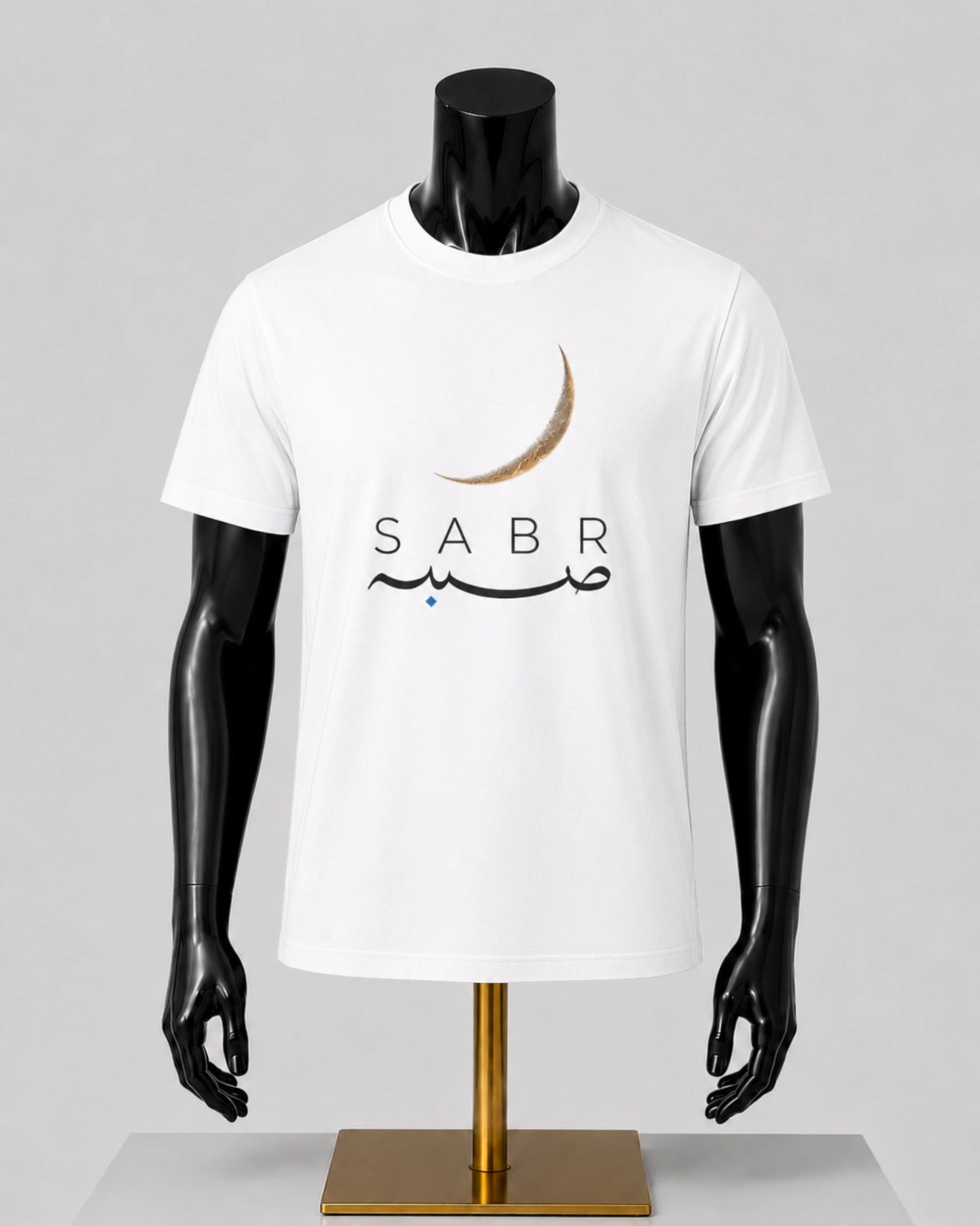 A white short-sleeve t-shirt displayed on a black mannequin torso with a gold stand, featuring a crescent moon and Arabic calligraphy with the word 'SABR' printed on the chest.