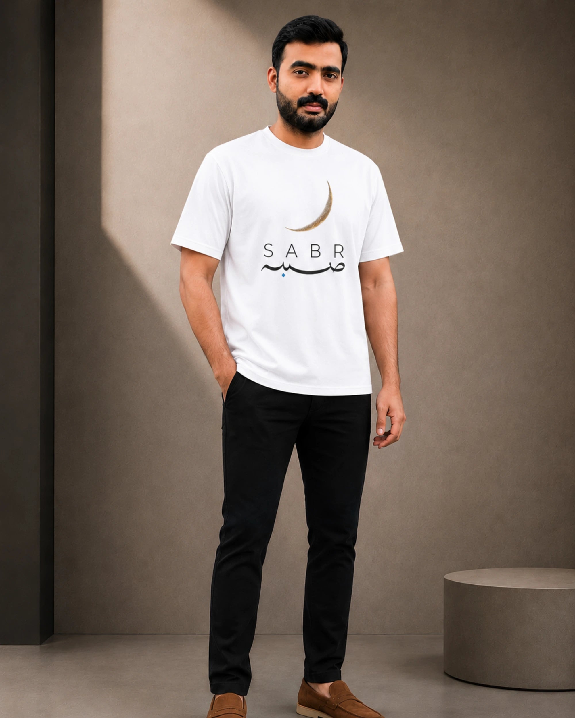A man wearing a white crew-neck t-shirt with a black and gold crescent moon and 'SABR' graphic, paired with black pants and brown loafers, standing in a minimalist studio setting.