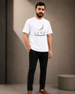 A man wearing a white crew-neck t-shirt with a black and gold crescent moon and 'SABR' graphic, paired with black pants and brown loafers, standing in a minimalist studio setting.