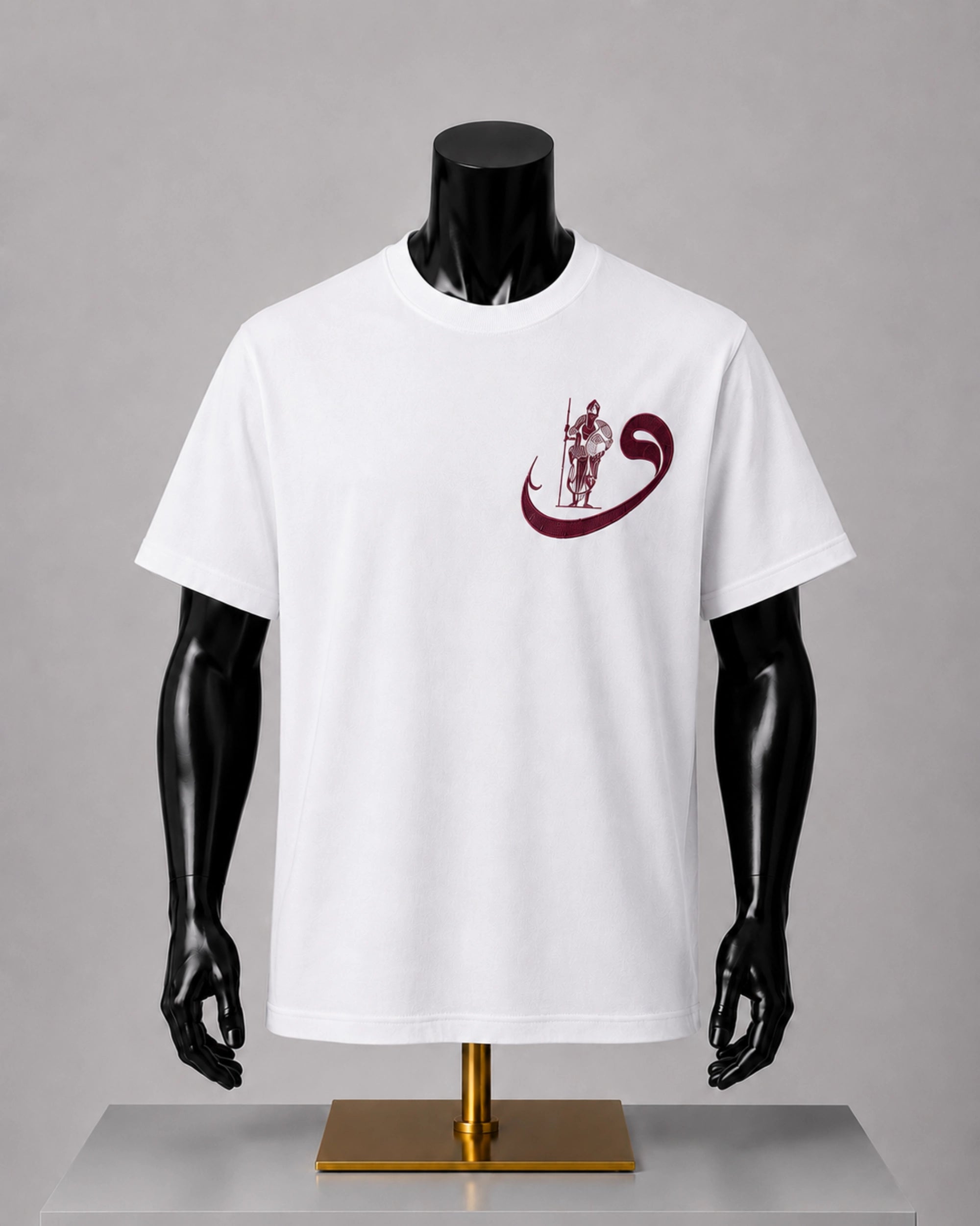 A white short-sleeve crewneck t-shirt displayed on a black mannequin torso with a gold stand against a neutral gray background. The shirt features a dark maroon graphic on the left chest depicting a warrior figure inside a crescent moon.