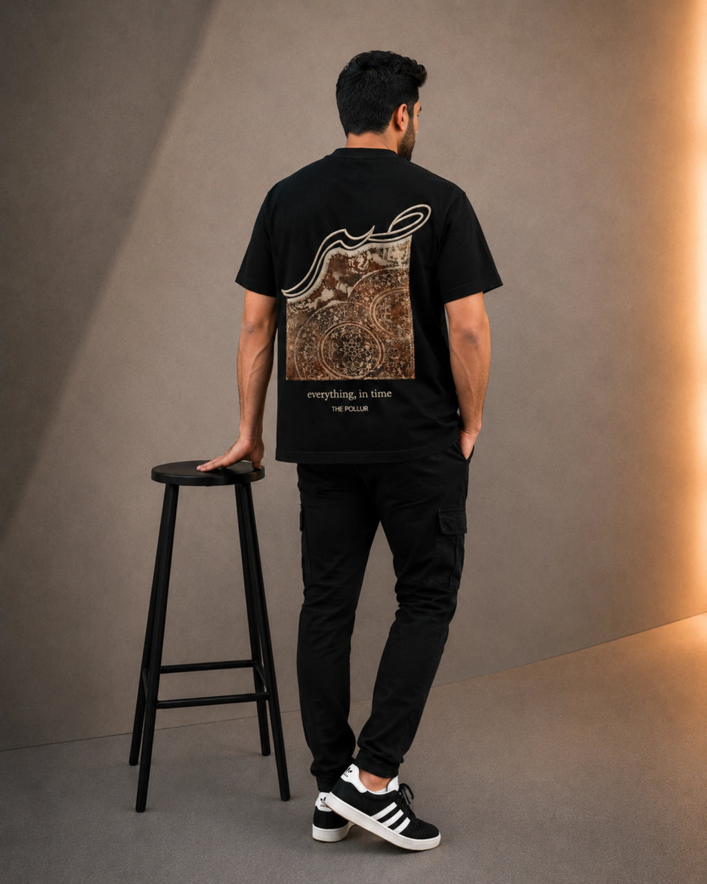 A man stands with his back to the camera wearing a black t-shirt with a large brown and white graphic print on the back, black cargo pants, and black and white sneakers. He is standing next to a black stool in a studio setting with a warm light on the right wall.