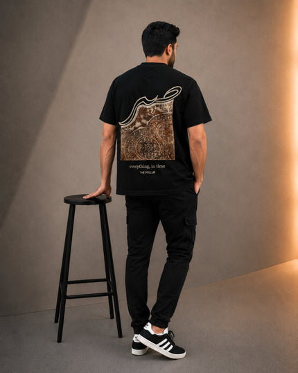 A man stands with his back to the camera wearing a black t-shirt with a large brown and white graphic print on the back, black cargo pants, and black and white sneakers. He is standing next to a black stool in a studio setting with a warm light on the right wall.