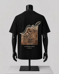 A black short-sleeve T-shirt displayed on a black mannequin torso with arms, shown from the back. The shirt features a large rectangular graphic print in brown and beige tones with an abstract white line above it, and the text 'everything, in time' and 'THE POLLUX' printed below the graphic.